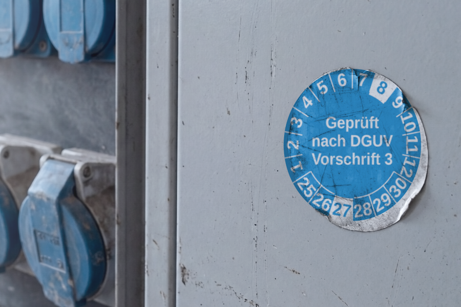 Abgenutzte DGUV-V3-Prüfplakette auf dem Blech eines Baustromverteilers, Monat 8 und Jahr 27 als nächster Prüftermin markiert, dahinter blaue CEE-Steckdosen