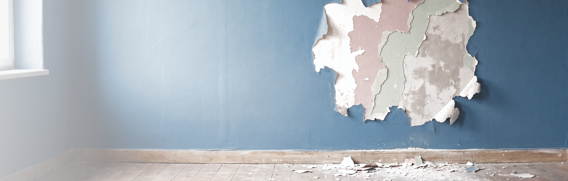 Leerer Altbauraum mit blau gestrichener Wand: rechts ist die Farbe auf einer großen Fläche abgeplatzt und gibt die älteren Anstriche darunter frei — creme, altrosa, salbeigrün und darunter der blanke Putz; auf den Dielen unter der Stelle liegen abgelöste Farbstücke